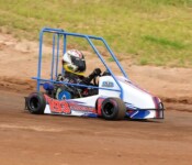A person driving a small, enclosed go-kart with the number 193 on a dirt track. The go-kart has a protective blue frame and the driver wears a helmet.