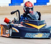 A person driving a racing go-kart on a track, wearing a helmet and gloves, with the go-kart featuring a "Pursuit" logo and number 99.