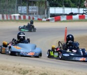 Two go-kart racers in black helmets compete on a dirt track, one in kart number 33 and the other in kart number 14. Other karts and a track fence are visible in the background.