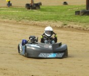 A driver in a white helmet races a go-kart on a dirt track surrounded by grass and tires.