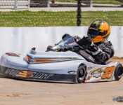 A racer in a go-kart with the number 33 on the front speeds around a track. The driver is wearing a helmet and protective gear, and the go-kart has a white body with the word "Pursuit" on it.