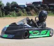 A person in black racing gear and helmet drives a small black and green go-kart with the number 95 on a dirt track, with stacks of tires visible in the background.