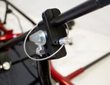 Close-up view of a mechanical component featuring a black clamp with a metal rod, bolt, and a wire loop. The component is attached to a red frame.