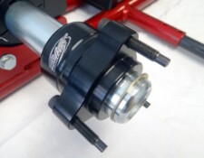 Close-up of a mechanical component with a black cylindrical housing, metallic interior parts, and protruding bolts, against a red metal frame background.